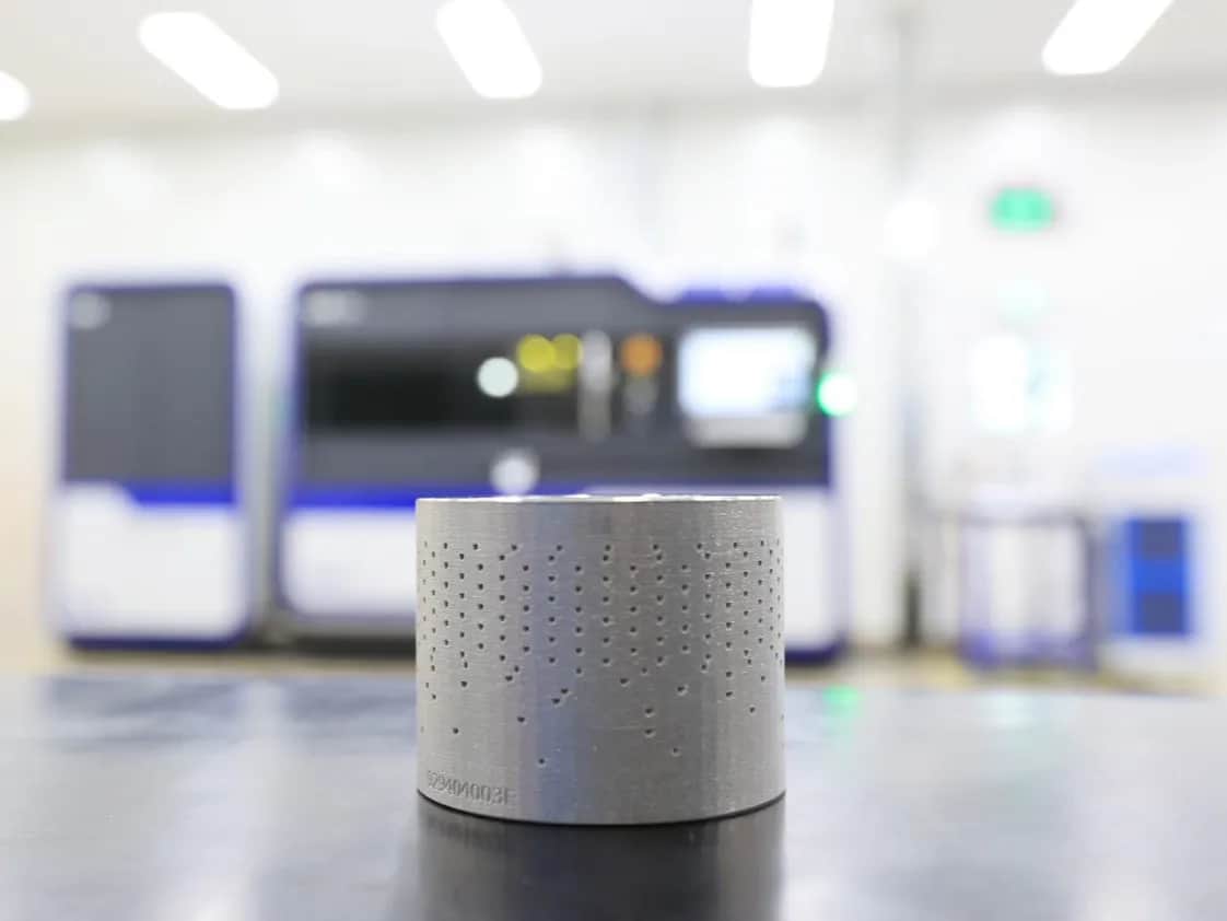 IMI CCI 3D-printed metal component in focus with manufacturing equipment in background.