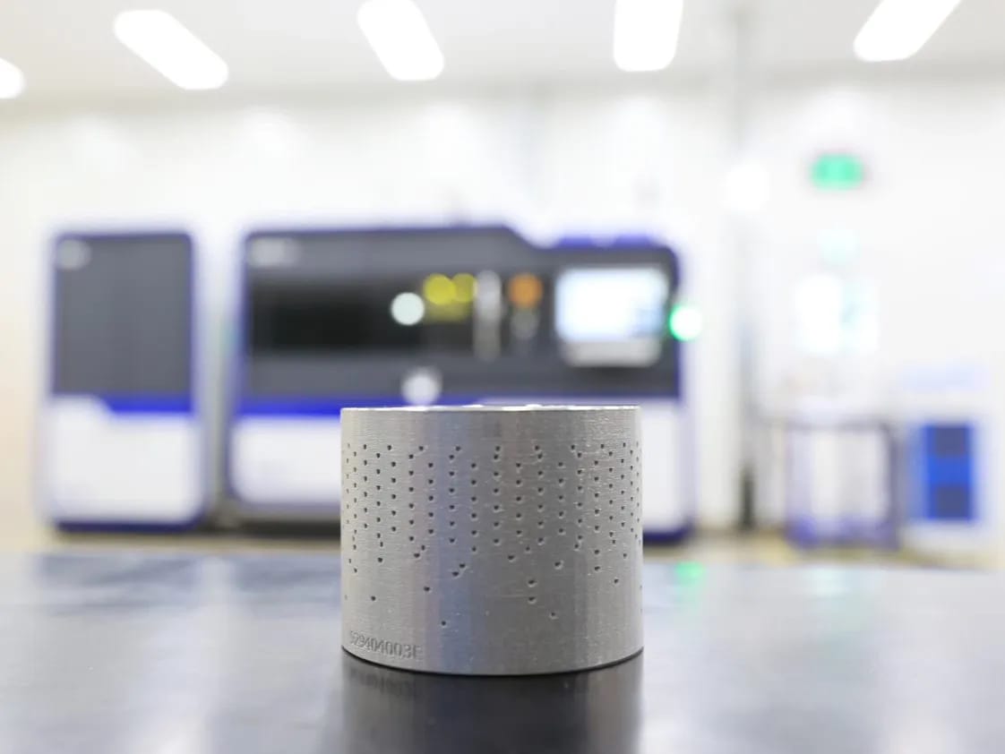 IMI CCI 3D-printed metal component in focus with manufacturing equipment in background.