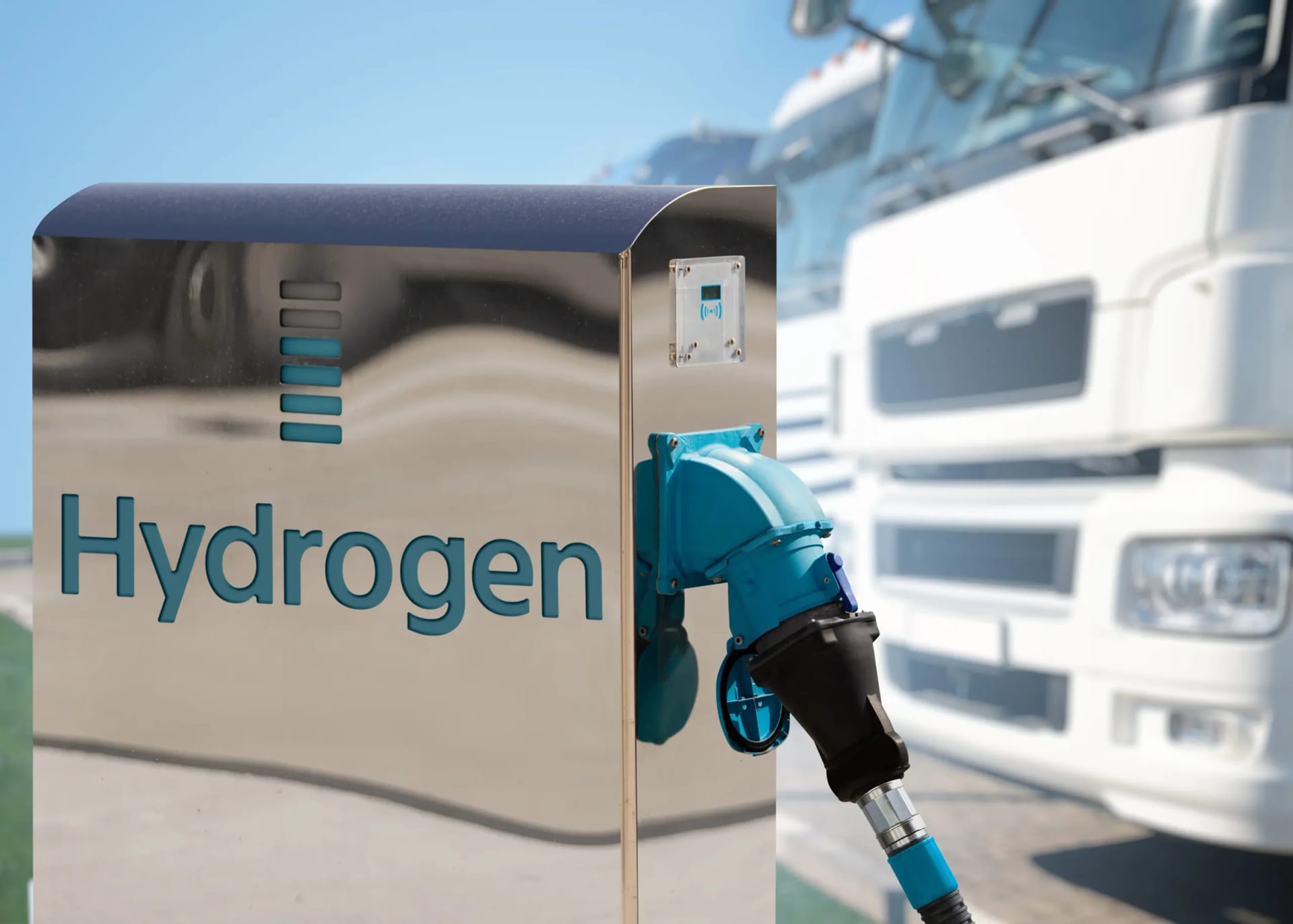 Hydrogen refueling station with metallic dispenser, blue writing, and trucks in background.