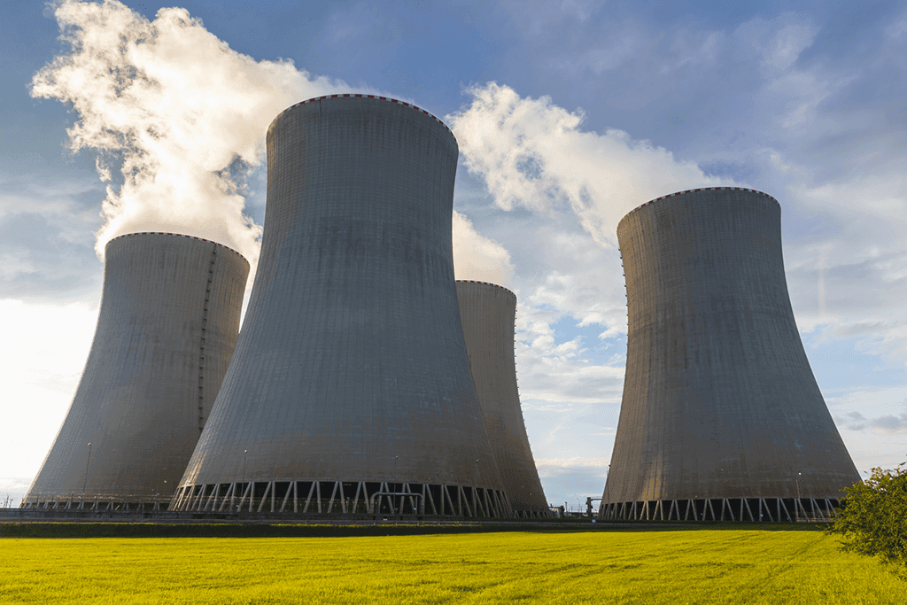 Four nuclear plant cooling towers emitting steam over a green field under a partly cloudy sky.