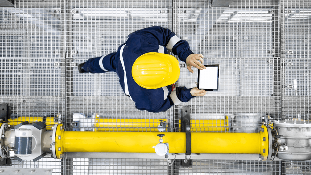 Engineer in safety gear uses a tablet above industrial piping system.