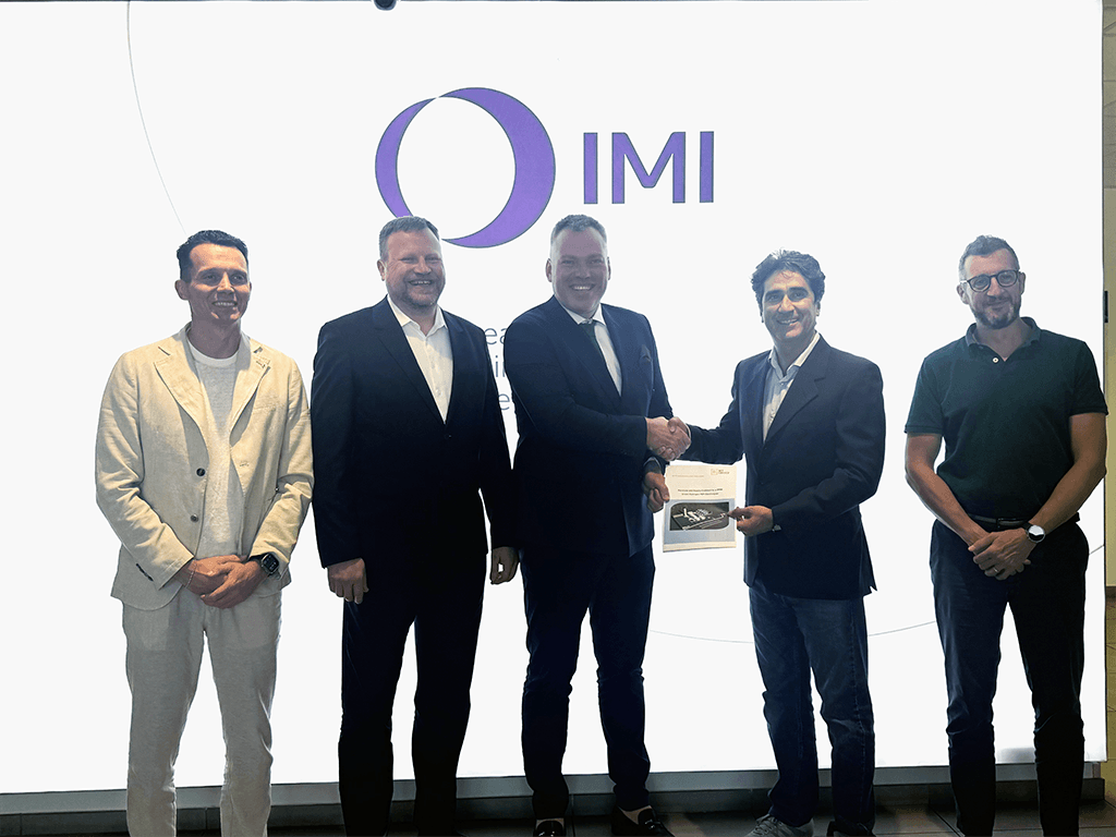 Five representatives from IMI, MT Group, and Vilnius District Heating stand in front of IMI branding; two shake hands holding a document.