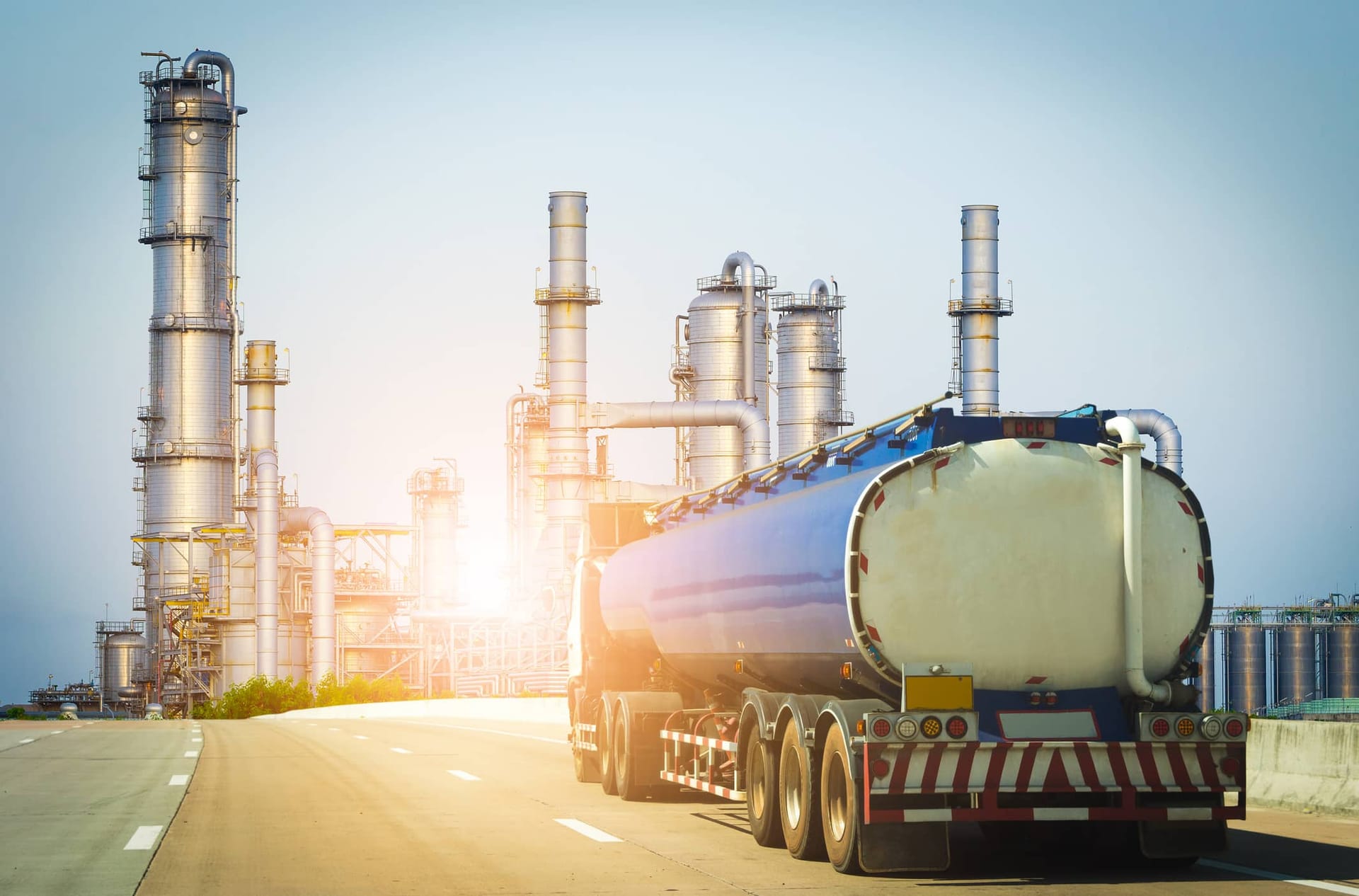 Tanker truck approaches large oil refinery complex with tall distillation towers at sunrise.