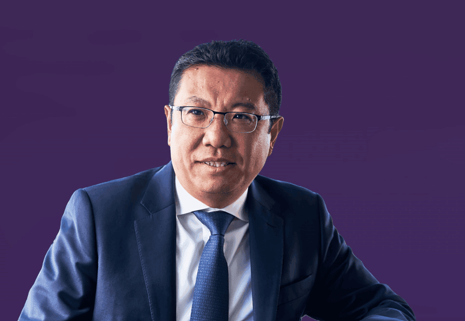 Person in dark blue suit, white shirt, and blue tie against a solid purple background. Face blurred.