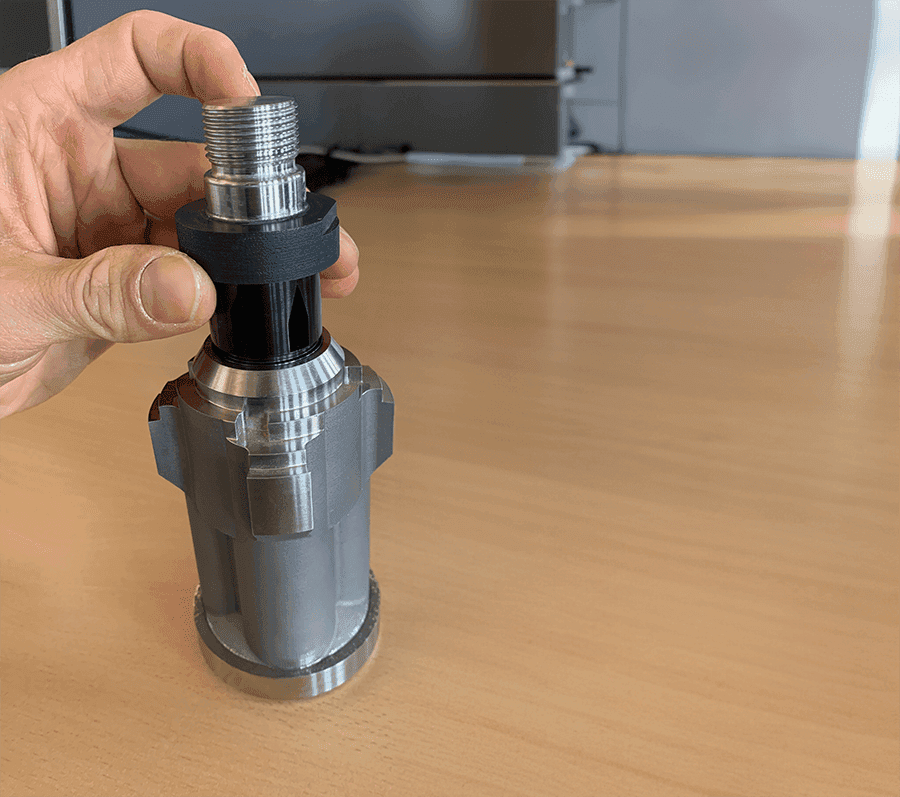 Hand holding metallic cylindrical component with threaded top, against wooden surface background.