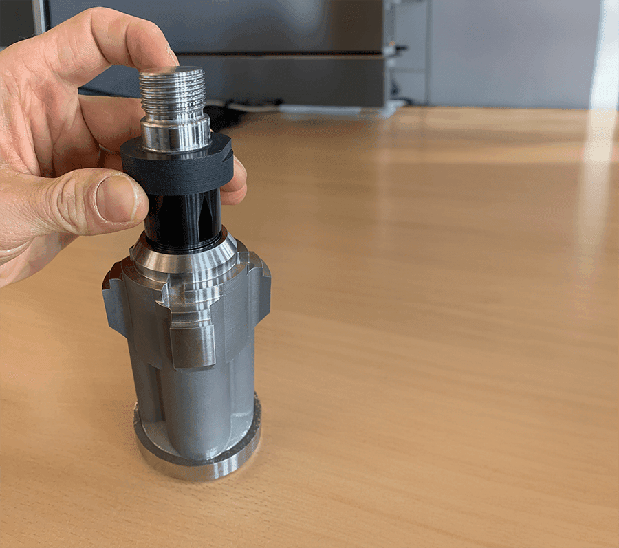 Hand holding metallic cylindrical component with threaded top, against wooden surface background.