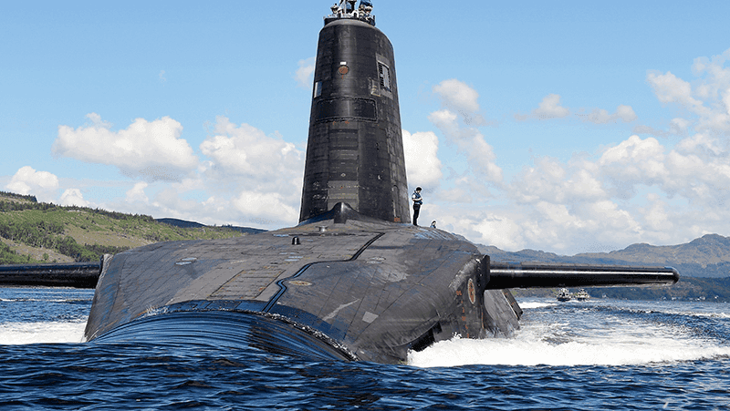 Submarine with visible conning tower partially submerged in open water near hilly coastline. Crown Copyright.