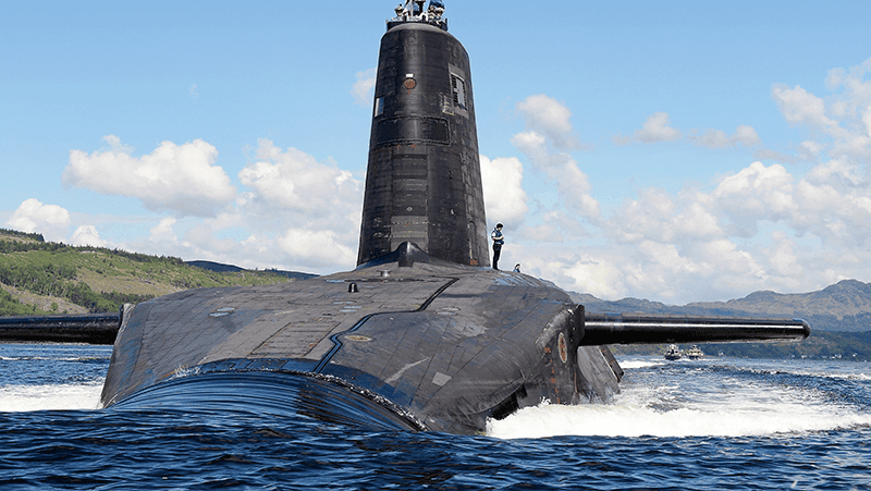 Submarine with visible conning tower partially submerged in open water near hilly coastline. Crown Copyright.